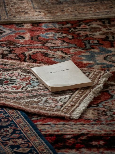 A worn floral-patterned rug with predominantly red, blue, and beige tones covers a wooden floor, displaying a slightly frayed edge. Placed on the rug is a small, white paper booklet titled 'images on 
