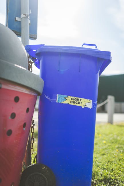 Five black plastic wheelie bins with yellow lids lined up against a plain, light gray wall in an outdoor setting. The ground is concrete, and each bin has a white label on the front with black text an