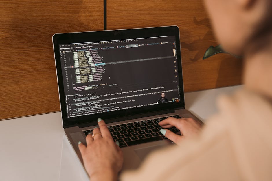 Close-up of a laptop keyboard and screen displaying lines of programming code, with the code editor showing syntax-highlighted text on a dark background. The laptop has a metallic body and black keys,